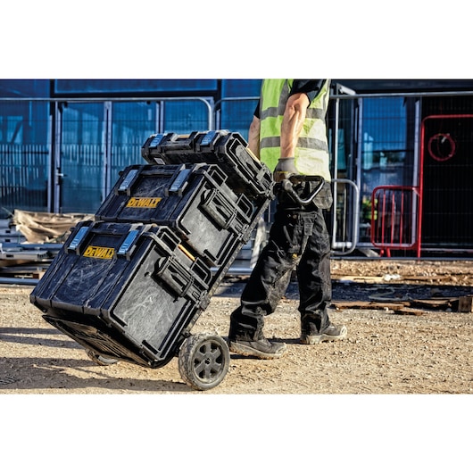 A person wearing work clothes and a high-visibility vest is pulling a stack of three DEWALT black toolboxes on a wheeled trolley at a construction site.
