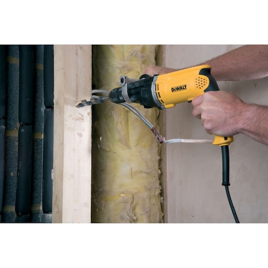 A person uses a yellow DEWALT electric drill to bore a hole through a wooden beam, with electrical cables and insulation visible in the background.