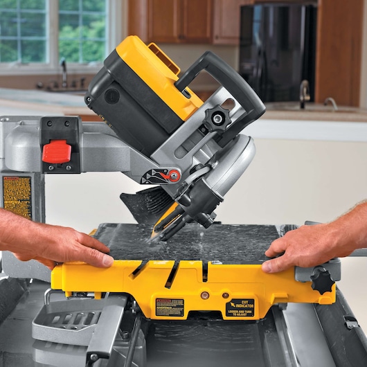 A close-up view of a DEWALT D24000 tile saw being used to cut a piece of tile. Two hands are guiding the tile under the saw's blade in a kitchen setting.