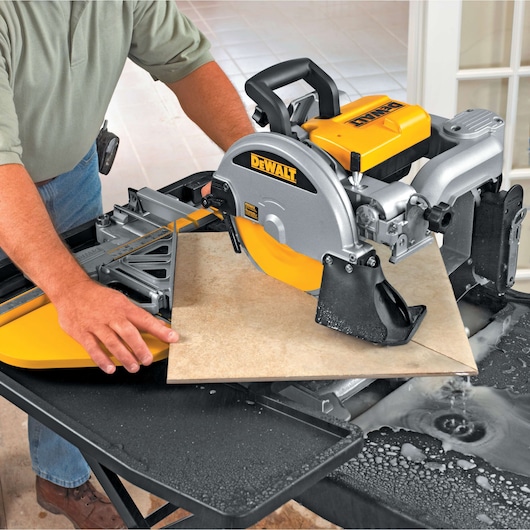 A person using a DEWALT D24000 tile saw to cut a ceramic tile on a worktable, with water helping to cool the blade and reduce dust.
