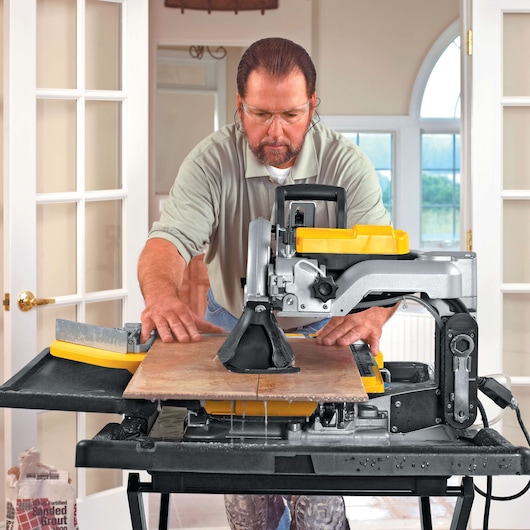 A person is using a DEWALT D24000 wet tile saw to cut a large tile indoors. The saw has a yellow and black design and is set up on a stand. Water is visible dripping onto the work surface. The person's face is blurred.