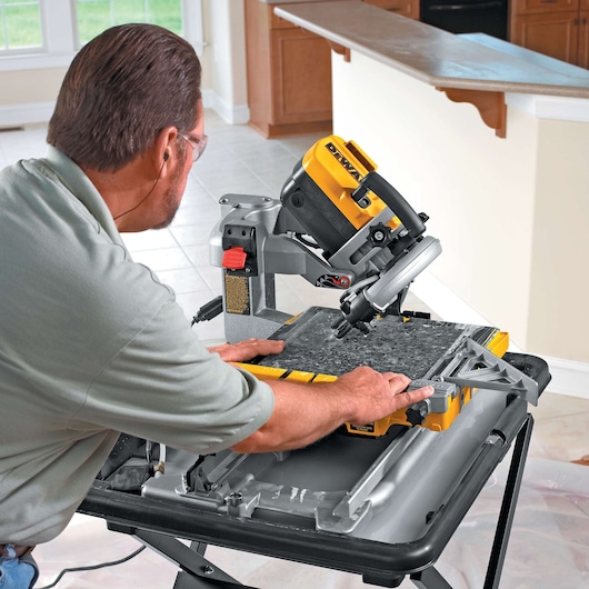 A person uses a DEWALT D24000 tile saw to cut a piece of stone or tile in a well-lit kitchen setting. The saw is mounted on a stand, and water is visible on the cutting surface.