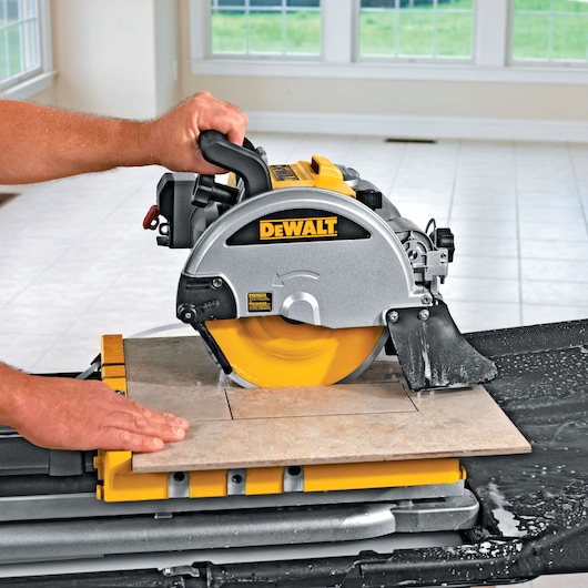A close-up view of a DEWALT tile saw in use, cutting a ceramic tile on a yellow workstation. Water is visible around the cutting area and the operator's hands are guiding the tile. The saw is being used in a bright tiled room with large windows.
