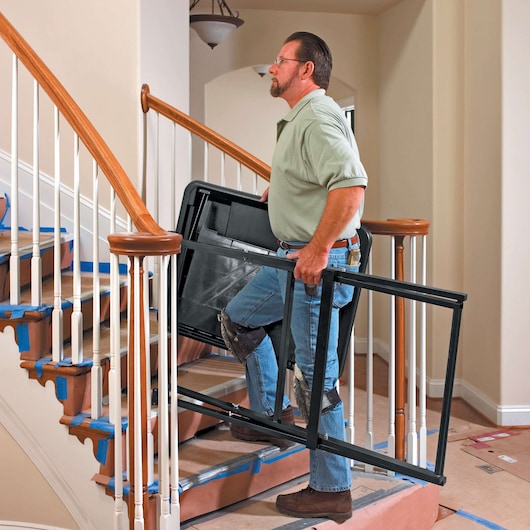 Person carrying DEWALT D24000 tile saw stand frame up a staircase with protective coverings on the steps.