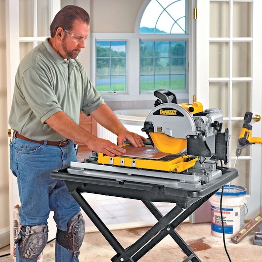 A person wearing knee pads is using a DEWALT tile saw (model D24000) on a stand in a well-lit room with large windows. Construction materials and tools are visible in the background.