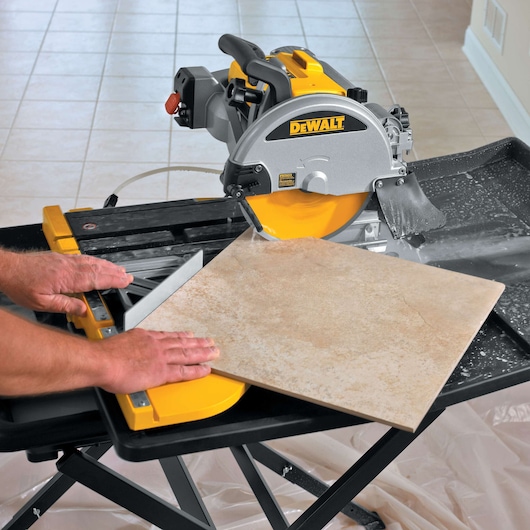 A DEWALT tile saw being used to cut a large ceramic tile. Water is flowing on the tile for cooling, and the tile is positioned on the saw's worktable with hands guiding it.
