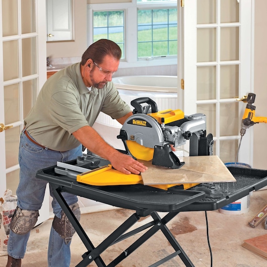 A person is using a DEWALT D24000 tile saw to cut a large ceramic tile in an indoor workspace. The saw is placed on a sturdy black stand, and water is visible on the table to cool the blade. The user's face is blurred for privacy, and they are wearing protective knee pads.