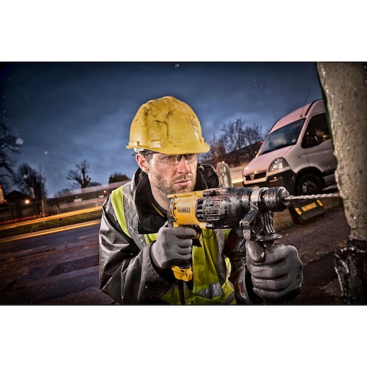 A construction worker wearing a yellow hard hat and reflective jacket uses a DEWALT hammer drill outdoors, with a white van and blurred lights in the background.