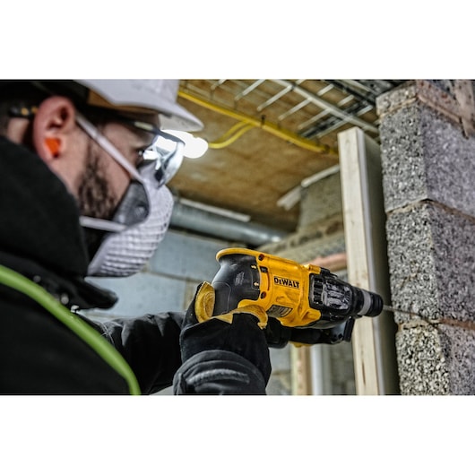 A worker wearing safety gear is using a yellow DEWALT hammer drill (model D25133K) to drill into a concrete wall at a construction site.