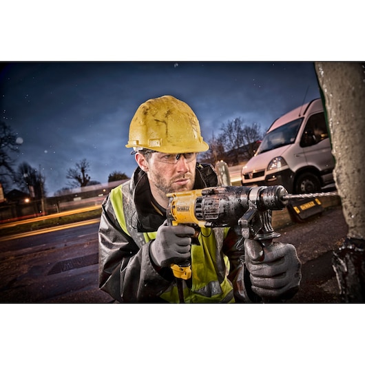 A construction worker wearing a yellow hard hat and reflective jacket uses a DEWALT hammer drill outdoors, with a white van and blurred lights in the background.