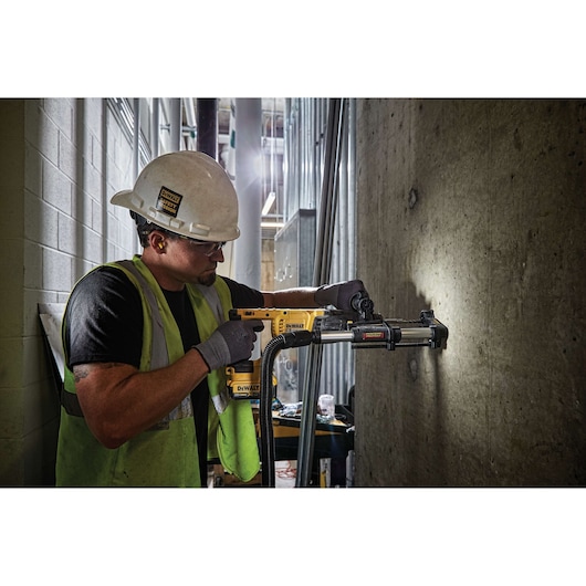A construction worker wearing a hard hat and safety vest operates a yellow DEWALT power tool with a dust extraction attachment, drilling into a concrete wall in an industrial setting.
