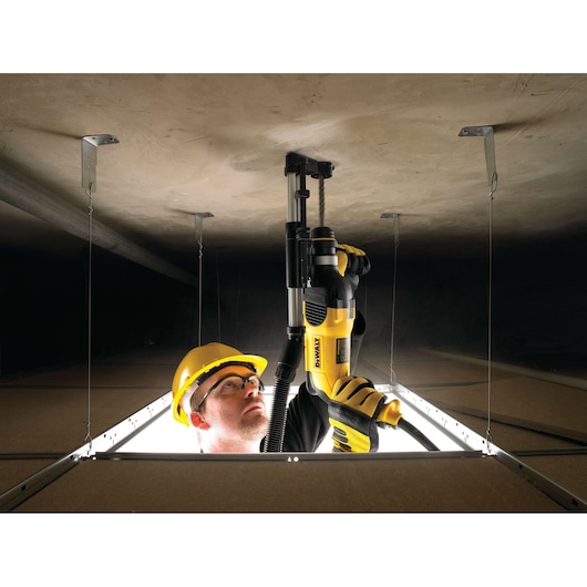 A person wearing safety gear and gloves uses a yellow DEWALT power tool to drill into a ceiling inside a construction site, viewed from an opening in the ceiling tiles.