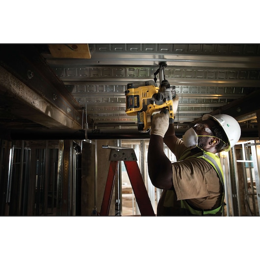 A construction worker wearing a hard hat, safety vest, gloves, and mask uses a DEWALT D25303DH tool to drill into a metal ceiling beam while standing on a ladder at a building site.