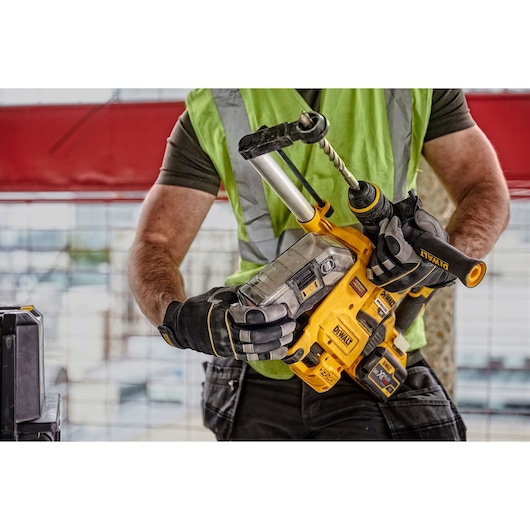 A person wearing a green safety vest and gloves is holding a DEWALT D25304DH cordless hammer drill with an attached dust extraction system on a construction site.