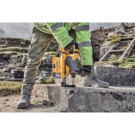 Person using DEWALT drill on concrete outdoors, wearing gloves, boots, and safety gear.