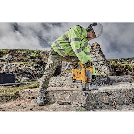 Worker drills into concrete outdoors using a yellow DEWALT tool, wearing safety gear and a high-visibility jacket.
