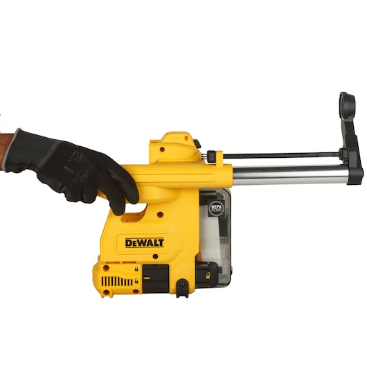 A person wearing a black glove is holding a yellow DEWALT dust extraction system attachment, model D25304DH, which has a HEPA filter and is designed for use with rotary hammers.