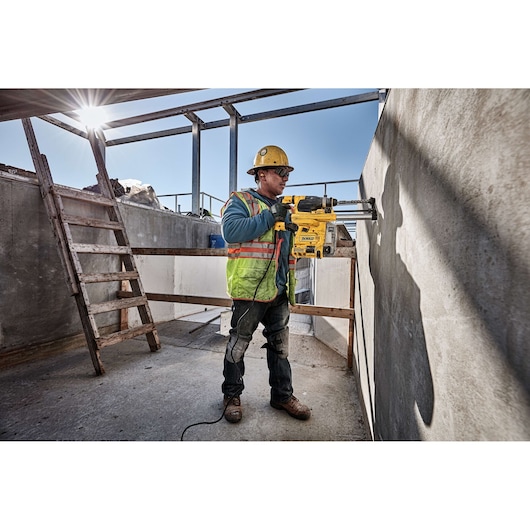 A construction worker wearing a safety helmet and high-visibility vest operates a yellow DEWALT rotary hammer drill, model D25333K, to bore a hole into a concrete wall at an outdoor construction site. The worker stands near a wooden ladder under a partly built structure with sunlight streaming in.