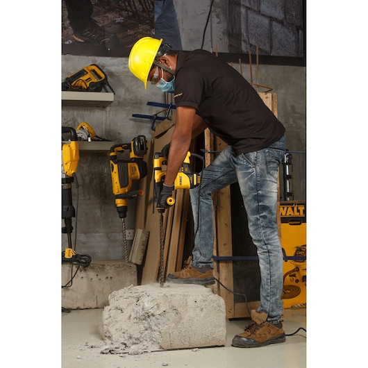 Worker with helmet using DEWALT rotary hammer on concrete block in industrial setting.