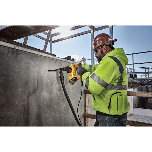 A person wearing a safety helmet and a high-visibility jacket is using a yellow DEWALT power tool to drill into a concrete wall at a construction site.