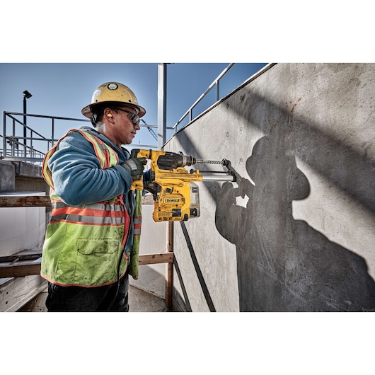 A construction worker wearing safety gear operates a DEWALT D25333K rotary hammer drill against a concrete wall at an outdoor job site.