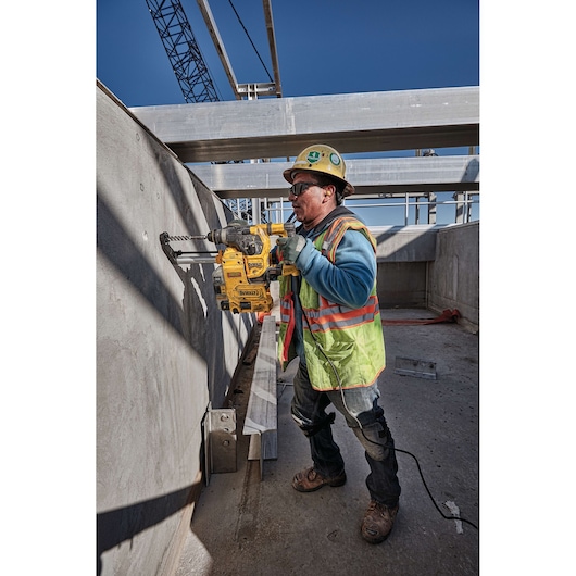 A construction worker wearing a safety vest and helmet is using a DEWALT D25333K rotary hammer drill to drill into a concrete wall at a construction site. The worker's face is blurred for privacy.