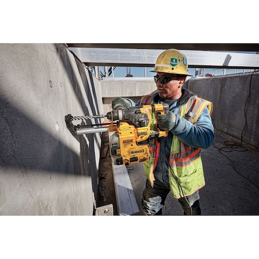 A construction worker wearing a safety vest and helmet is using a yellow DEWALT rotary hammer drill (model D25333K) to drill holes into a concrete wall at a construction site.