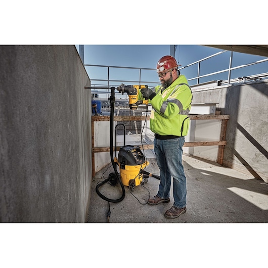 A construction worker wearing a red hard hat and high-visibility jacket is using a yellow DEWALT power tool to drill into a concrete wall on a job site. A yellow vacuum device is connected to the tool to collect dust.