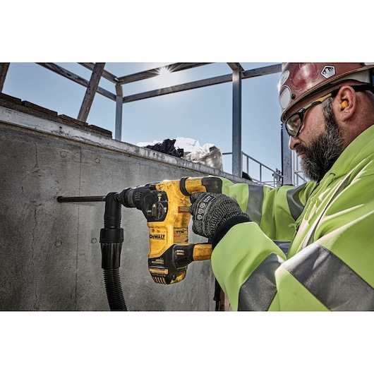 A person wearing safety gear is using a yellow DEWALT power drill to make a hole in a concrete wall at a construction site. The person's face is not visible clearly.