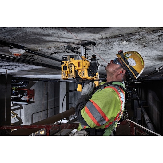 A construction worker wearing safety gear operates a DEWALT rotary hammer drill to bore a hole in a concrete ceiling. The worker is elevated on a platform in an industrial setting.