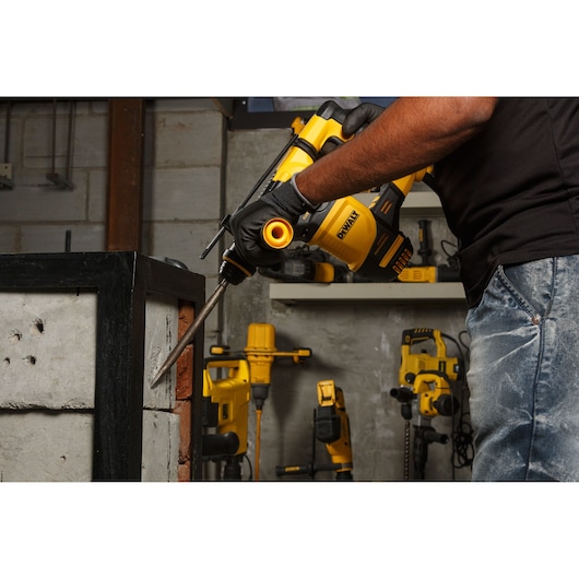 Person uses a DEWALT hammer drill on a concrete and brick wall in a workshop with more tools visible in the background.
