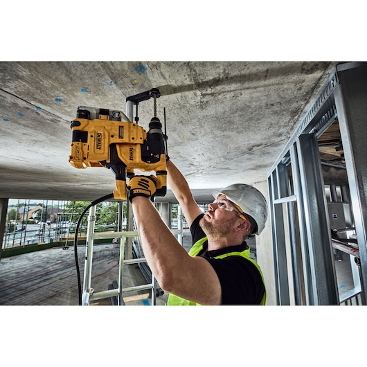 A person wearing a safety helmet and vest is using a DEWALT power tool to drill into a concrete ceiling at a construction site. The DEWALT tool is held overhead and appears to be a rotary hammer drill.