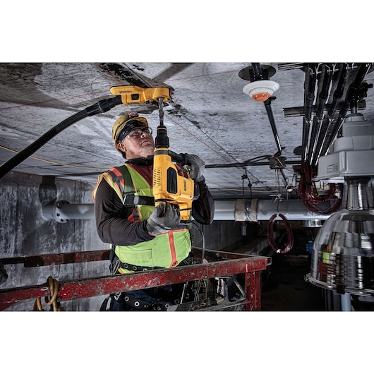 Worker uses a DEWALT drill on a ceiling at a construction site, wearing safety gear and a high-visibility vest.