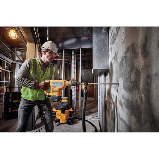 A person wearing safety gear and a helmet operates a DEWALT D25614K rotary hammer drill against a concrete wall in a construction site. DEWALT equipment and tools are visible in the background.
