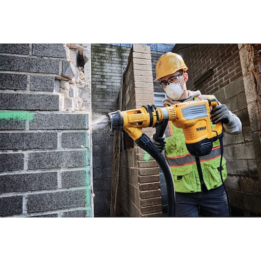 A construction worker wearing a yellow hard hat and safety vest uses a DEWALT rotary hammer drill (model D25614K) to break through a brick wall, with dust being expelled through an attached dust extraction system.