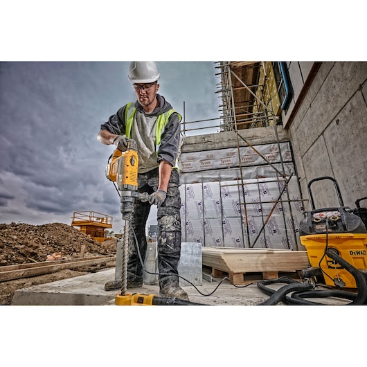 A construction worker wearing a safety helmet and high-visibility vest operates a DEWALT rotary hammer drill (SKU: D25733K) on a building site. The worker's face is blurred, and there is construction equipment, scaffolding, and insulation materials visible in the background.