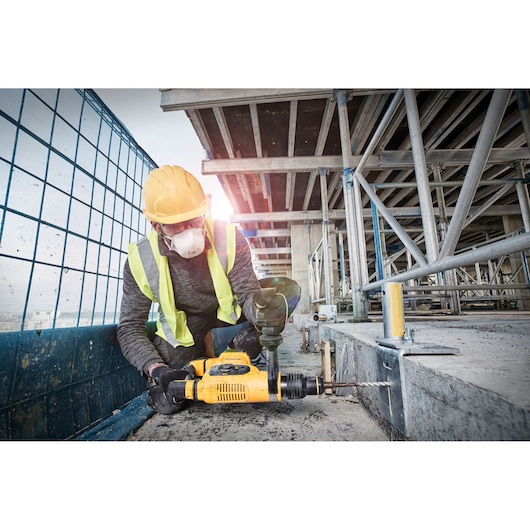 A construction worker wearing a yellow hard hat and reflective vest is using a yellow DEWALT hammer drill to drill into a concrete surface at a building site.