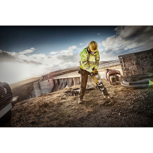A construction worker wearing a yellow hard hat and high-visibility jacket uses a DEWALT jackhammer on rocky ground near an industrial structure outdoors, with equipment and vehicles visible around.