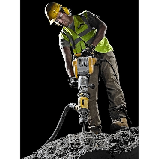 A construction worker wearing a yellow helmet and a high-visibility vest operates a DEWALT demolition hammer on a rough concrete surface. The worker's face is blurred for privacy, and the background is black, highlighting the tool and worker.
