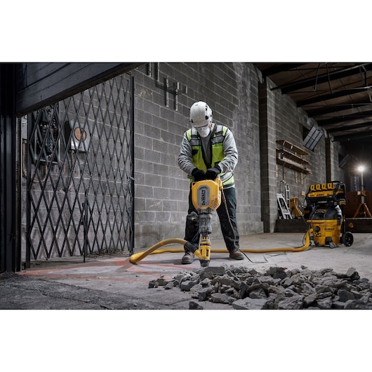 A worker wearing a safety helmet and high-visibility vest is using a yellow DEWALT jackhammer to break up concrete inside an