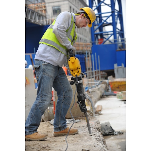 Worker uses a DEWALT D25960K demolition hammer on concrete at a construction site, wearing safety gear.