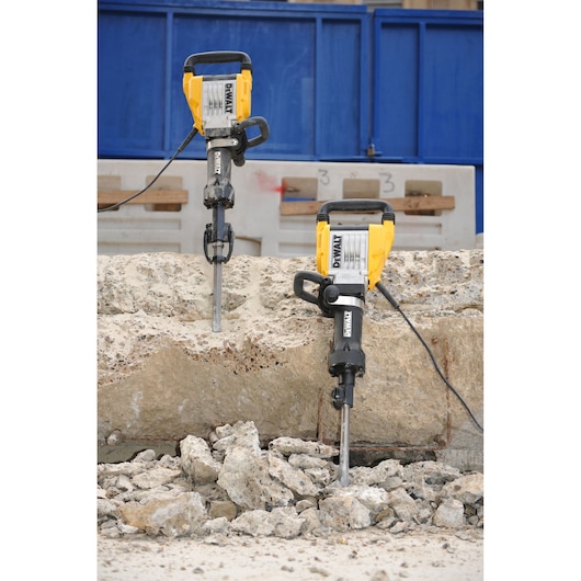 Two DEWALT demolition hammers are positioned upright on a large concrete block at a construction site, surrounded by broken pieces of concrete.