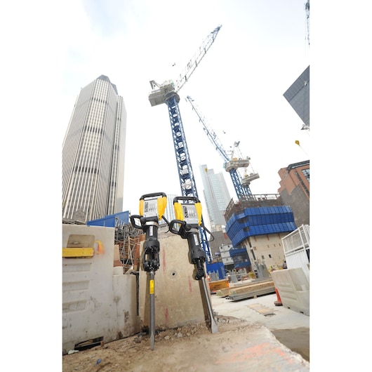 Two DEWALT D25960K demolition hammers are positioned upright at a construction site with tall buildings and cranes in the background.