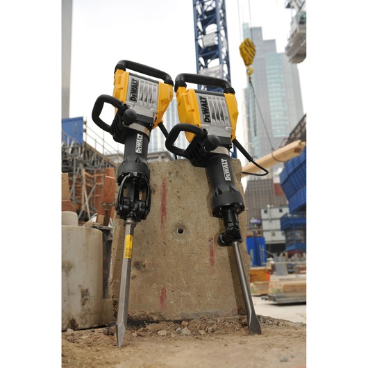 Two DEWALT electric demolition hammers are positioned upright on a concrete block at a construction site, with buildings and cranes visible in the background.