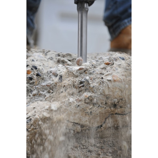 A close-up of a demolition hammer breaking apart concrete, with dust and debris falling. The tool is being operated outdoors.