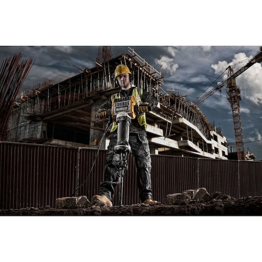 A construction worker using a DEWALT D25981 demolition hammer on a construction site, with a partially built structure and cranes in the background.