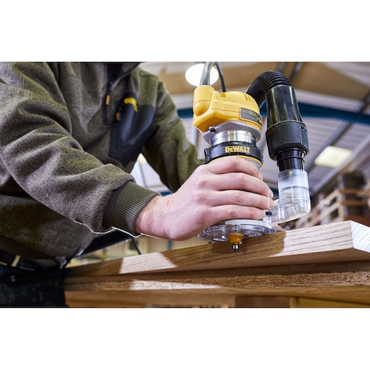 A person is using a DEWALT router tool, model D26200, to shape wood in a workshop. The router has a dust extraction attachment connected.