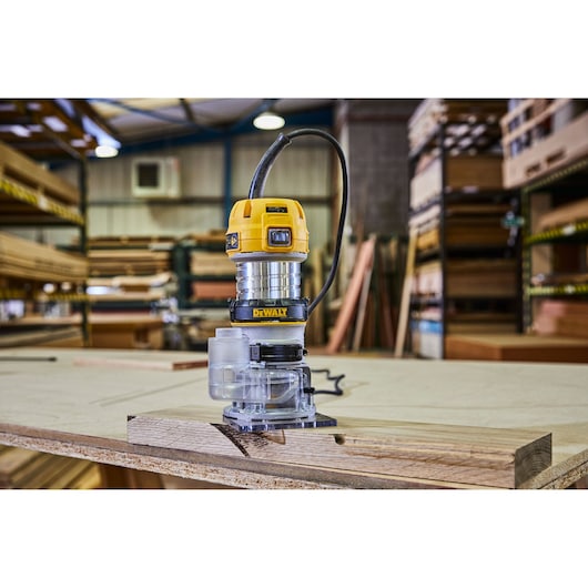 A DEWALT electric router model D26200 is placed on a piece of wood in a workshop. The tool is yellow and silver with a cord attached, and woodworking materials are visible in the background.