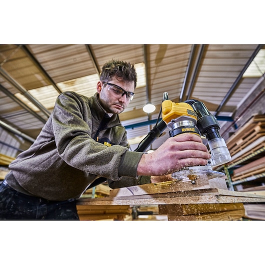 A person using a DEWALT D26200 router tool to work on a piece of wood in a workshop. The individual's face is blurred and there are wood shavings visible during the operation.