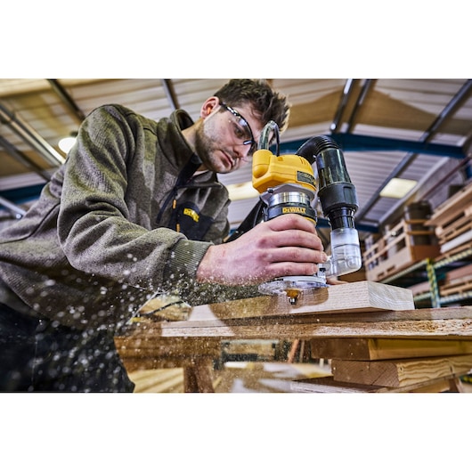 A person using a DEWALT D26200 router on a piece of wood, with sawdust flying, in a woodworking workshop.